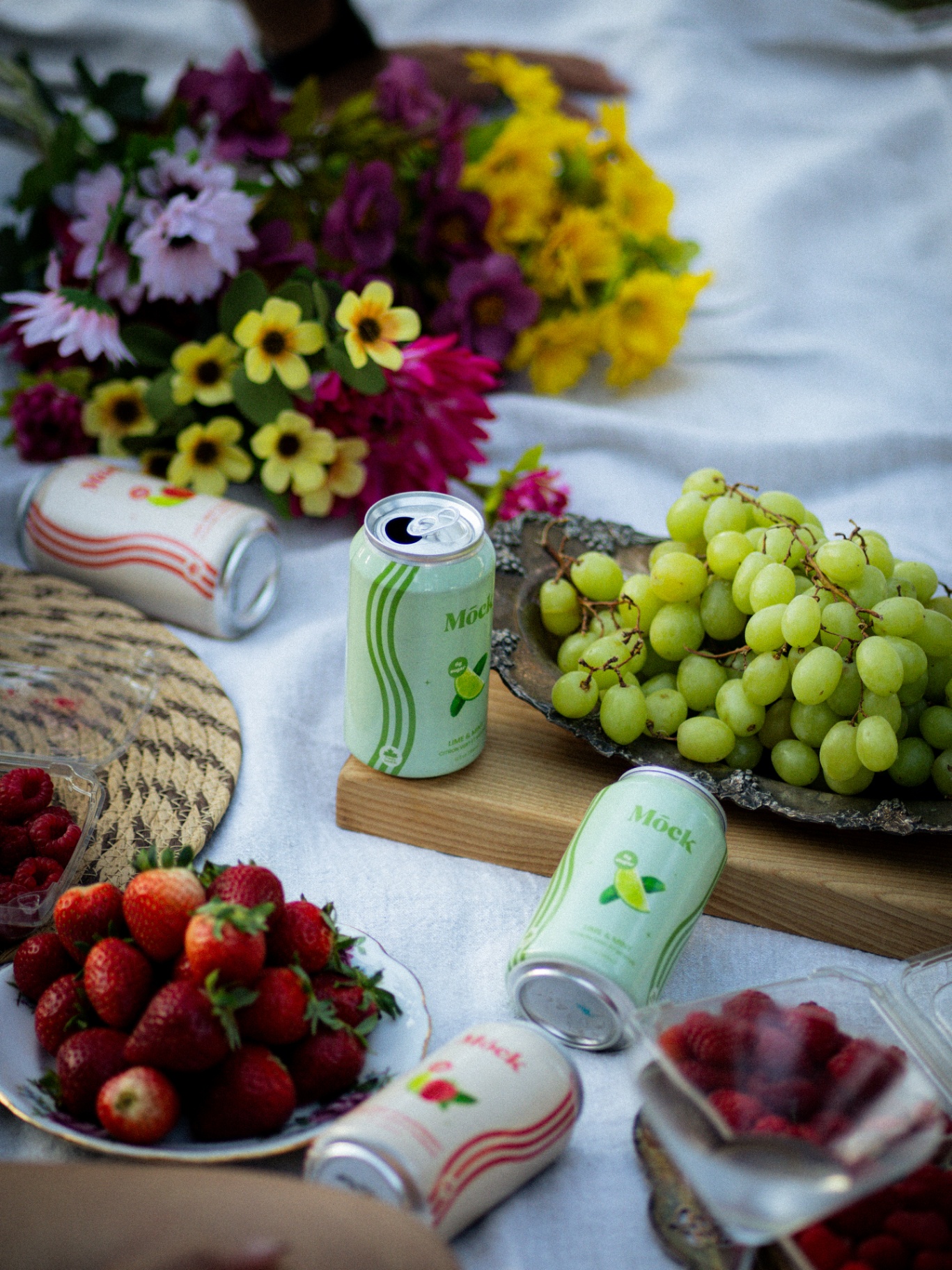 Mōck cans on a picnic blanket with flowers, grapes, and berries